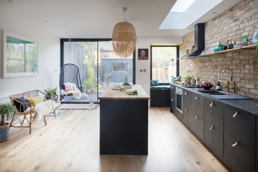 Mid-sized scandinavian single-wall kitchen in London with a double-bowl sink, flat-panel cabinets, black cabinets, wood benchtops, beige splashback, brick splashback, stainless steel appliances, light hardwood floors, with island and beige floor.