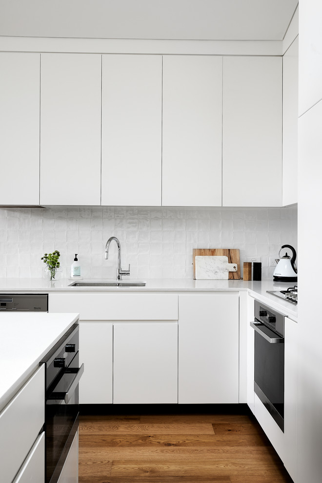 Photo of a contemporary l-shaped kitchen in Sydney with an undermount sink, flat-panel cabinets, white cabinets, white splashback, black appliances, medium hardwood floors, with island, brown floor and white benchtop.