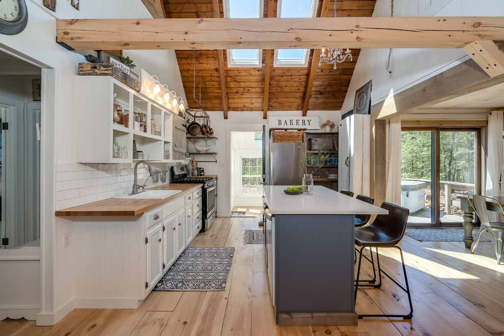 This is an example of a country eat-in kitchen in Portland Maine with a drop-in sink, open cabinets, white cabinets, wood benchtops, white splashback, subway tile splashback, stainless steel appliances, light hardwood floors and with island.