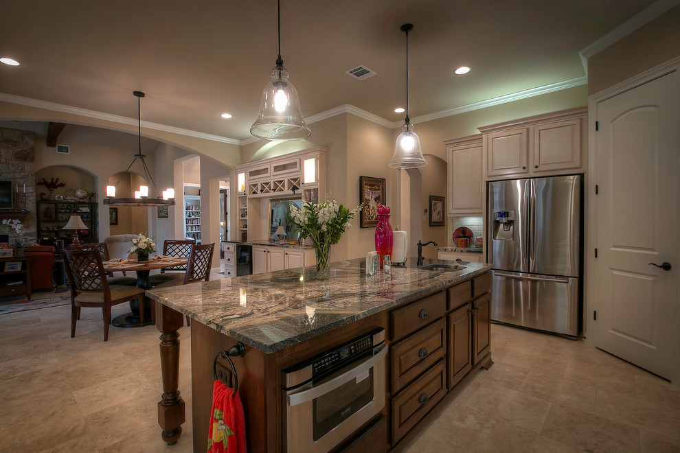 This is an example of a country eat-in kitchen in Austin with an undermount sink, raised-panel cabinets, white cabinets, granite benchtops, beige splashback, travertine splashback, stainless steel appliances, travertine floors, with island, beige floor and black benchtop.