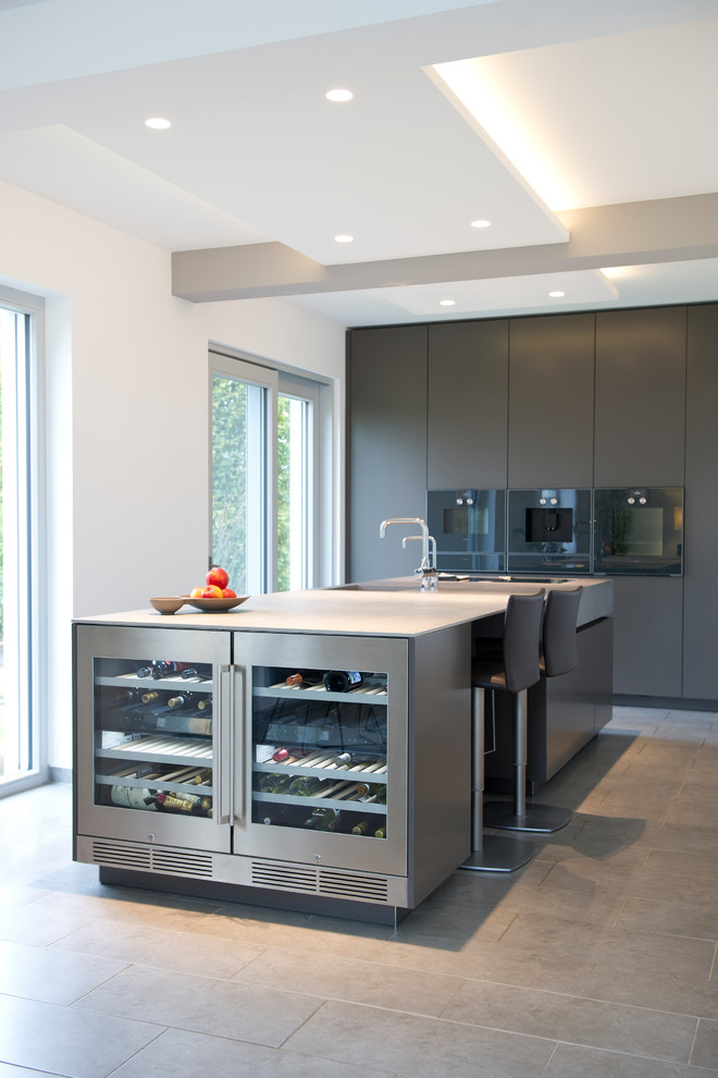 This is an example of a large contemporary galley open plan kitchen in Other with an integrated sink, flat-panel cabinets, grey cabinets, stainless steel benchtops, black appliances, cement tiles, with island, grey floor, white splashback and white benchtop.