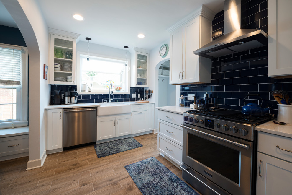 Mid-sized country u-shaped eat-in kitchen in Indianapolis with a farmhouse sink, shaker cabinets, white cabinets, quartz benchtops, blue splashback, subway tile splashback, stainless steel appliances, porcelain floors, no island and brown floor.