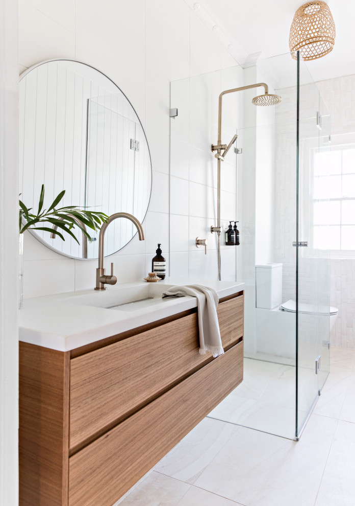 This is an example of a contemporary 3/4 bathroom in Sydney with flat-panel cabinets, medium wood cabinets, a curbless shower, a two-piece toilet, white tile, an integrated sink, white floor, a hinged shower door and white benchtops.