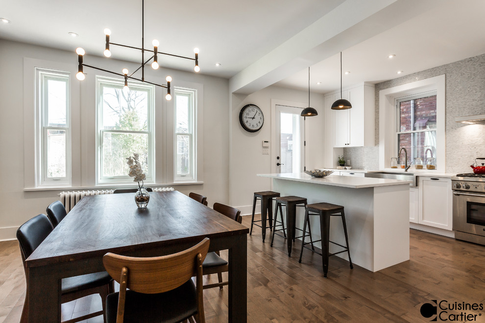 Mid-sized transitional l-shaped medium tone wood floor eat-in kitchen photo in Montreal with a farmhouse sink, shaker cabinets, white cabinets, quartzite countertops, white backsplash, mosaic tile backsplash, stainless steel appliances and an island