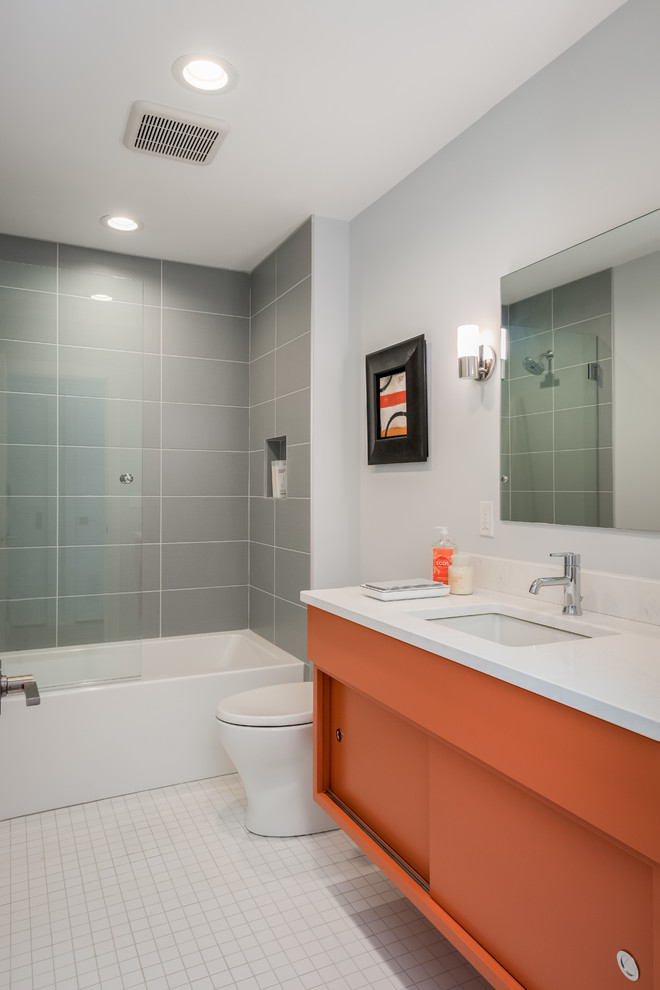 Mid-sized midcentury kids bathroom in New York with flat-panel cabinets, orange cabinets, an alcove tub, a shower/bathtub combo, gray tile, ceramic tile, grey walls, ceramic floors, an undermount sink, engineered quartz benchtops, white floor and white benchtops.