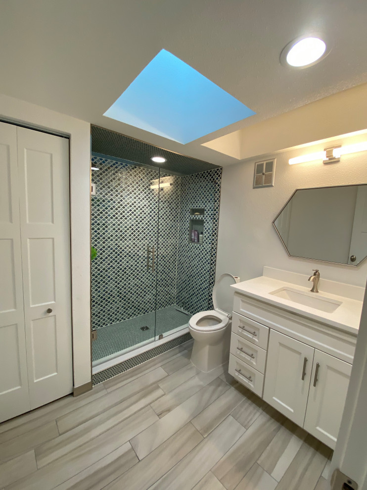 This is an example of a small beach style master bathroom in Tampa with shaker cabinets, white cabinets, an alcove shower, a two-piece toilet, multi-coloured tile, glass tile, blue walls, porcelain floors, an undermount sink, engineered quartz benchtops, beige floor, a hinged shower door and white benchtops.
