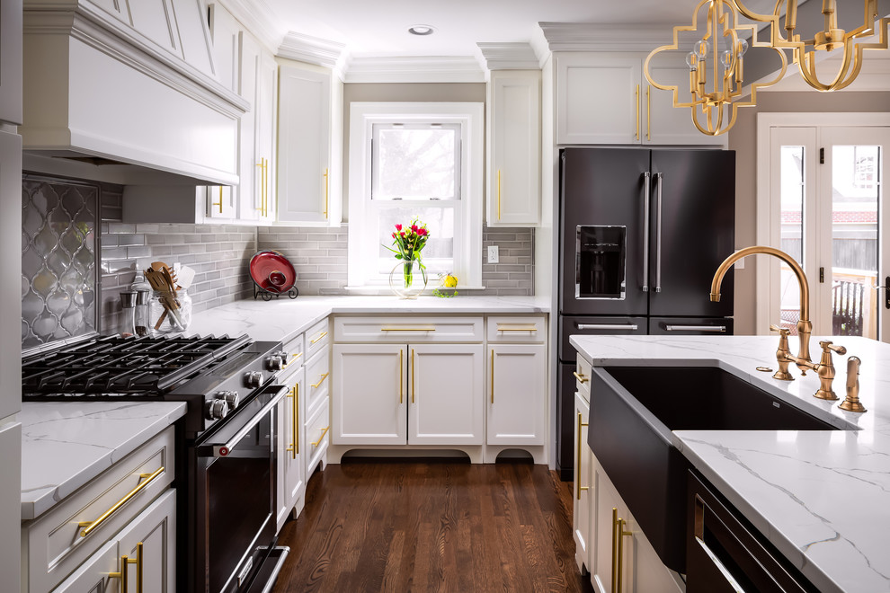 Example of a transitional l-shaped medium tone wood floor and brown floor kitchen design with a farmhouse sink, recessed-panel cabinets, white cabinets, gray backsplash, black appliances, an island and white countertops