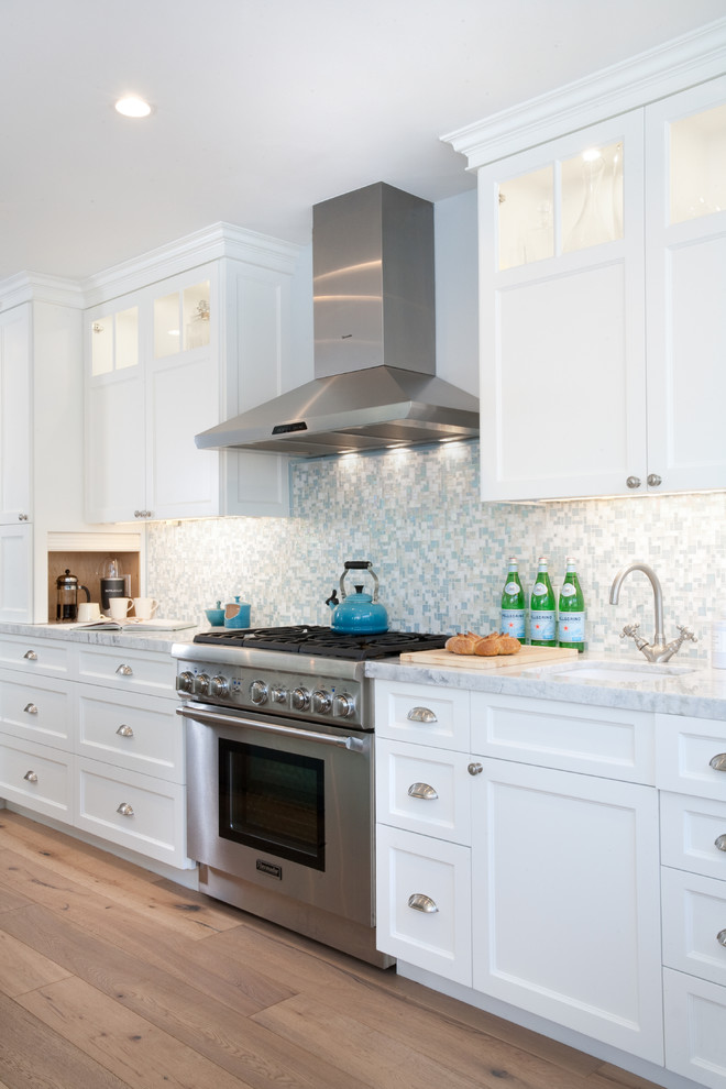 Inspiration for a large beach style l-shaped eat-in kitchen in Los Angeles with a farmhouse sink, shaker cabinets, white cabinets, blue splashback, mosaic tile splashback, stainless steel appliances, medium hardwood floors, with island and marble benchtops.