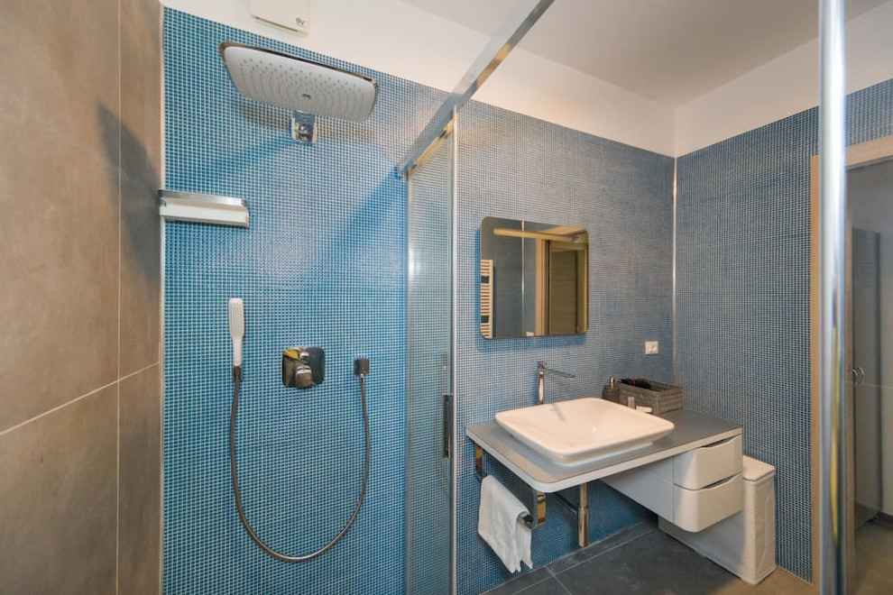 Photo of a mid-sized modern 3/4 bathroom in Other with open cabinets, white cabinets, a curbless shower, a wall-mount toilet, blue tile, mosaic tile, white walls, porcelain floors, a vessel sink, stainless steel benchtops, grey floor, a sliding shower screen and grey benchtops.