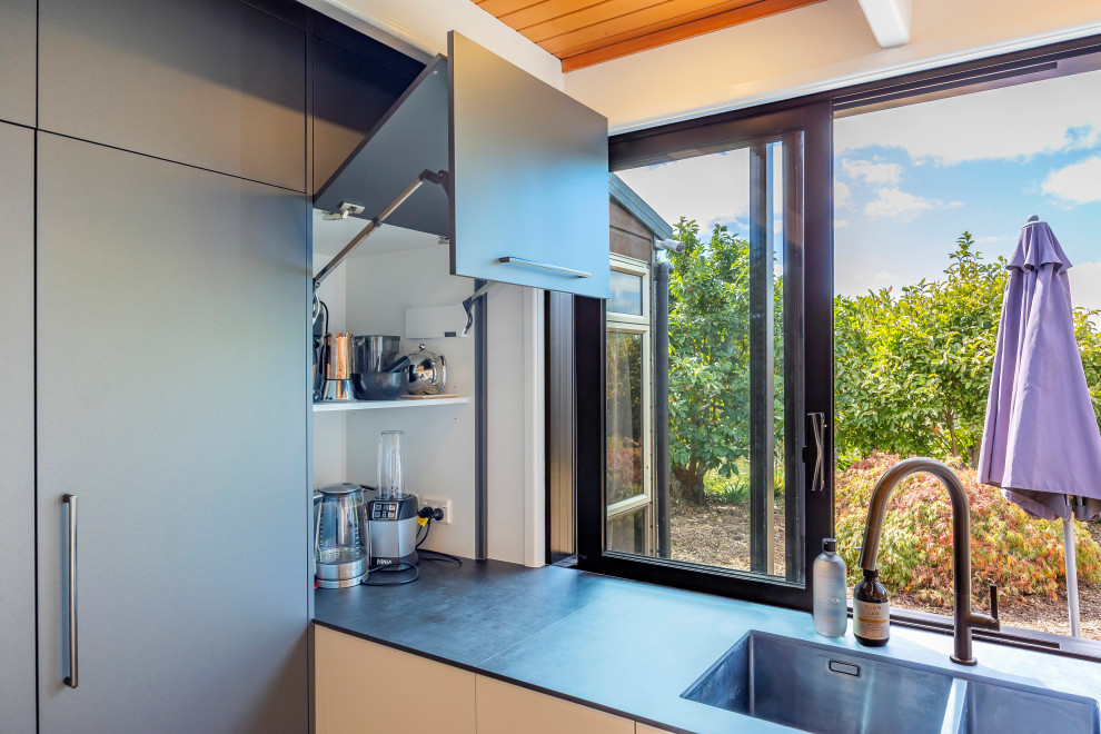 Design ideas for a large modern l-shaped eat-in kitchen in Auckland with an undermount sink, flat-panel cabinets, grey cabinets, white splashback, black appliances, laminate floors, with island, brown floor, grey benchtop and exposed beam.