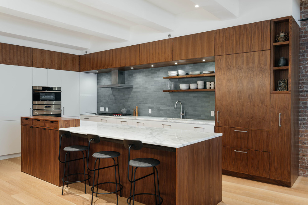 Photo of a contemporary l-shaped kitchen in New York with an undermount sink, flat-panel cabinets, dark wood cabinets, grey splashback, subway tile splashback, panelled appliances, light hardwood floors, with island and white benchtop.