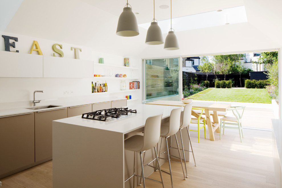 Design ideas for a mid-sized contemporary l-shaped eat-in kitchen in London with flat-panel cabinets, solid surface benchtops, white splashback, with island, an undermount sink, beige cabinets, light hardwood floors, glass sheet splashback, stainless steel appliances, beige floor and beige benchtop.