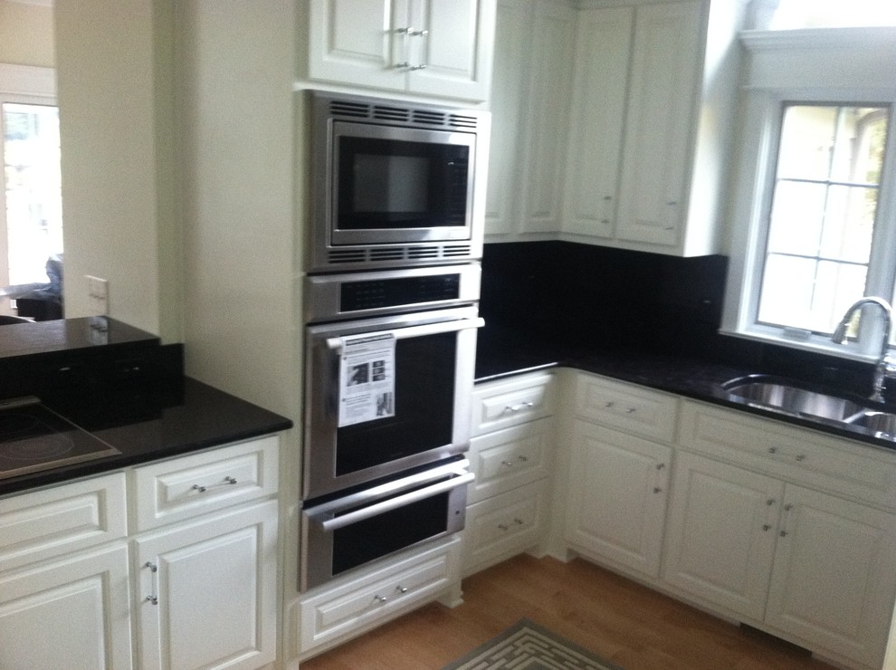 Transitional u-shaped light wood floor kitchen photo with raised-panel cabinets, white cabinets, granite countertops, black backsplash, stone slab backsplash, black appliances and no island