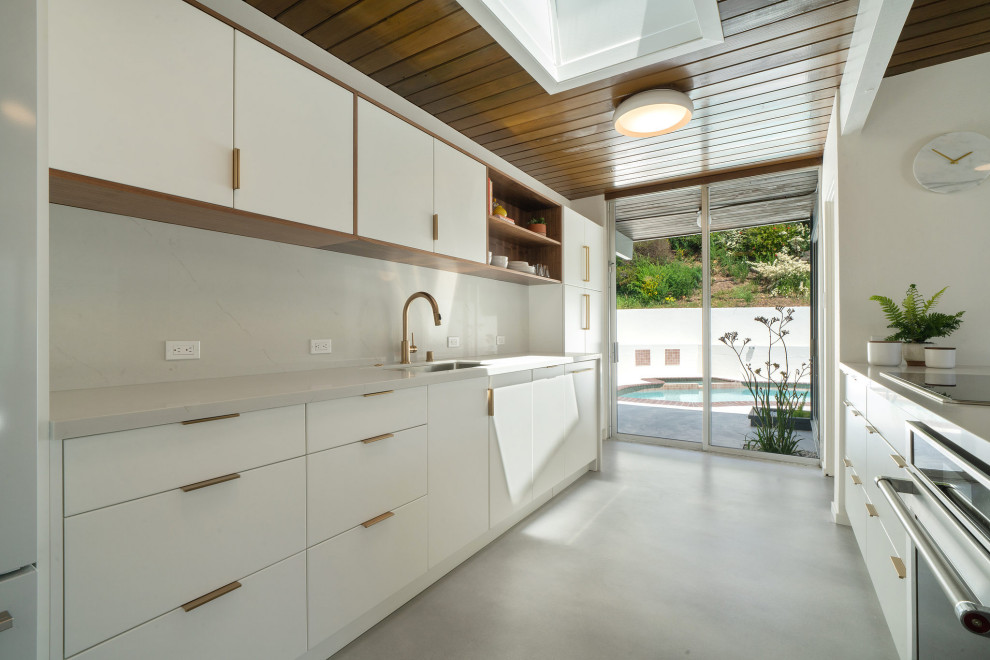 This is an example of a midcentury galley kitchen in San Francisco with an undermount sink, flat-panel cabinets, white cabinets, stainless steel appliances, concrete floors, grey floor, white benchtop and wood.