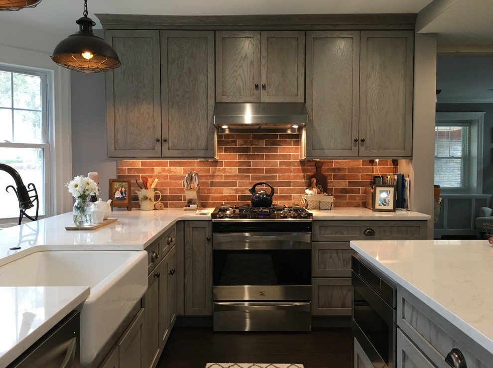 Photo of a large country l-shaped eat-in kitchen in Philadelphia with a farmhouse sink, shaker cabinets, grey cabinets, quartz benchtops, red splashback, brick splashback, stainless steel appliances, dark hardwood floors, with island, brown floor and white benchtop.
