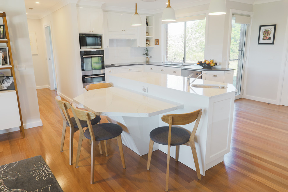 This is an example of a large transitional galley eat-in kitchen in Sydney with a double-bowl sink, flat-panel cabinets, white cabinets, quartz benchtops, white splashback, ceramic splashback, stainless steel appliances, light hardwood floors, with island, brown floor and white benchtop.