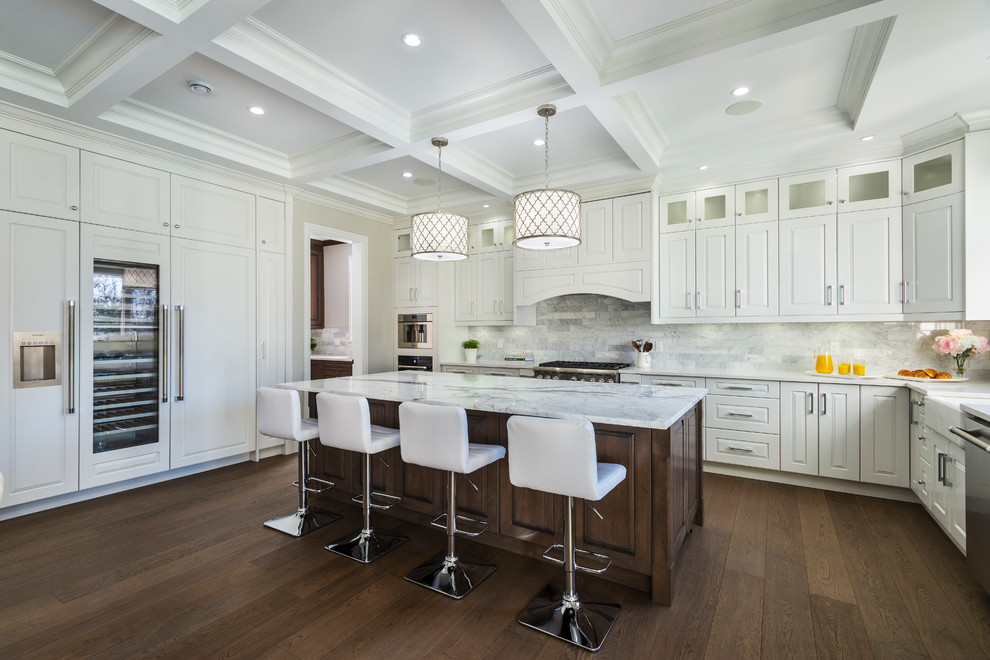 This is an example of an expansive traditional u-shaped open plan kitchen in Vancouver with a farmhouse sink, raised-panel cabinets, white cabinets, quartzite benchtops, grey splashback, marble splashback, stainless steel appliances, medium hardwood floors, with island, brown floor and grey benchtop.