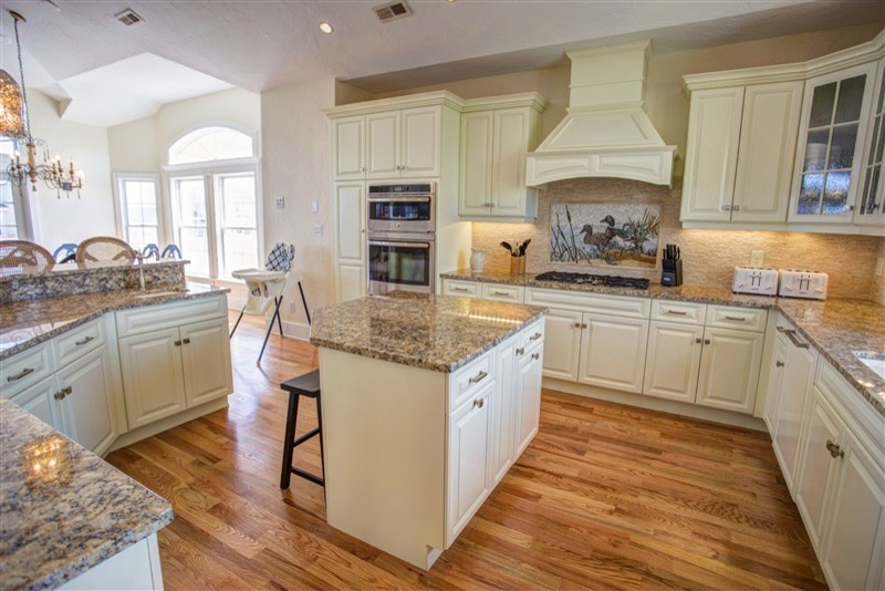 Open concept kitchen - mid-sized transitional l-shaped medium tone wood floor open concept kitchen idea with an undermount sink, raised-panel cabinets, beige cabinets, granite countertops, beige backsplash, stone tile backsplash, stainless steel appliances and an island