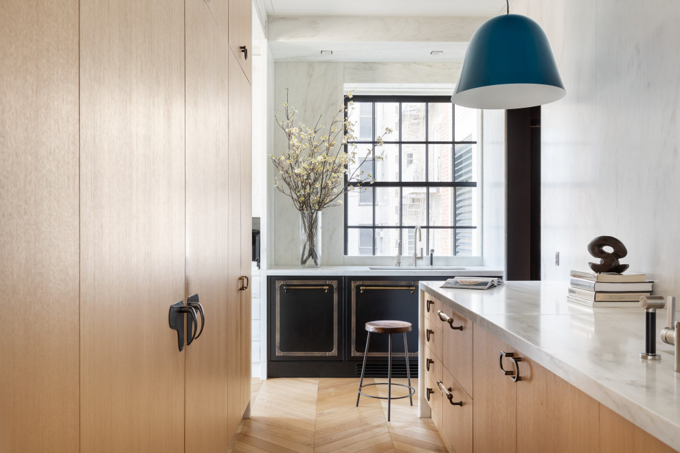 This is an example of a mid-sized eclectic galley open plan kitchen in New York with an integrated sink, light wood cabinets, marble benchtops, white splashback, marble splashback, black appliances, light hardwood floors, with island, brown floor and white benchtop.