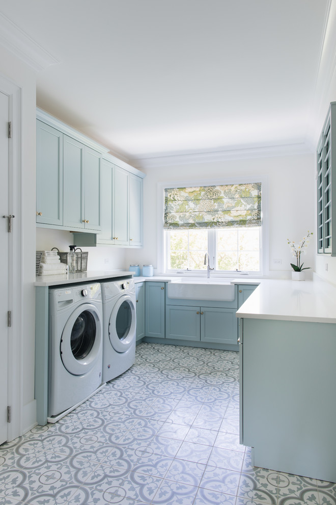 This is an example of a beach style u-shaped dedicated laundry room in Chicago with a farmhouse sink, shaker cabinets, blue cabinets, white walls, a side-by-side washer and dryer, multi-coloured floor and white benchtop.