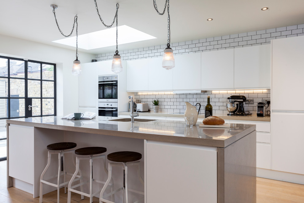 This is an example of a contemporary kitchen in London with flat-panel cabinets, white splashback, subway tile splashback, light hardwood floors and with island.