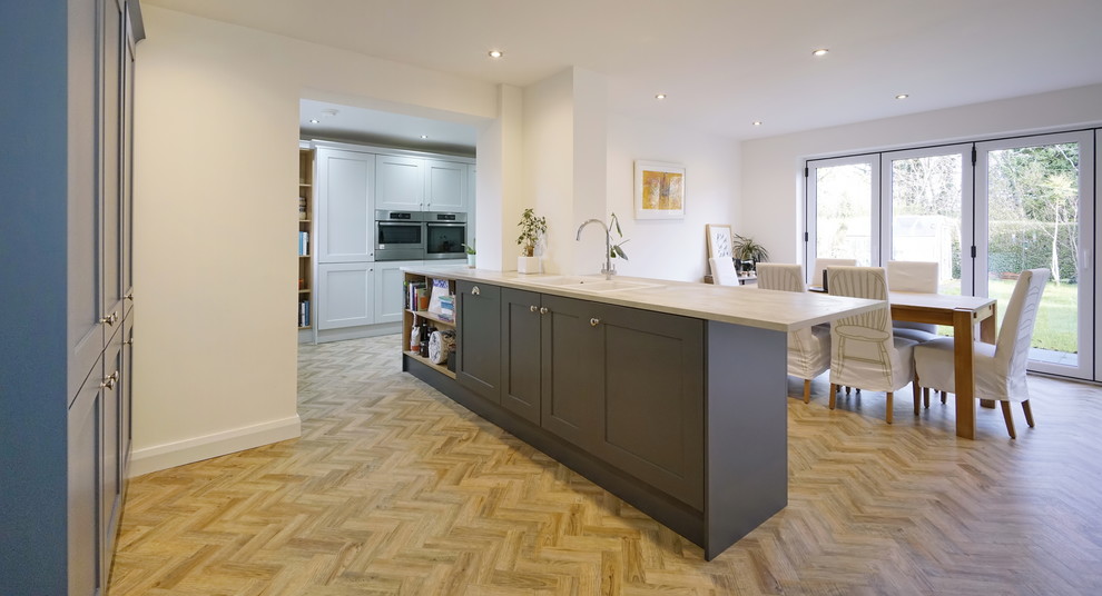 This is an example of a large contemporary l-shaped open plan kitchen in Belfast with a double-bowl sink, shaker cabinets, grey cabinets, laminate benchtops, white splashback, ceramic splashback, panelled appliances, vinyl floors, a peninsula, brown floor and grey benchtop.