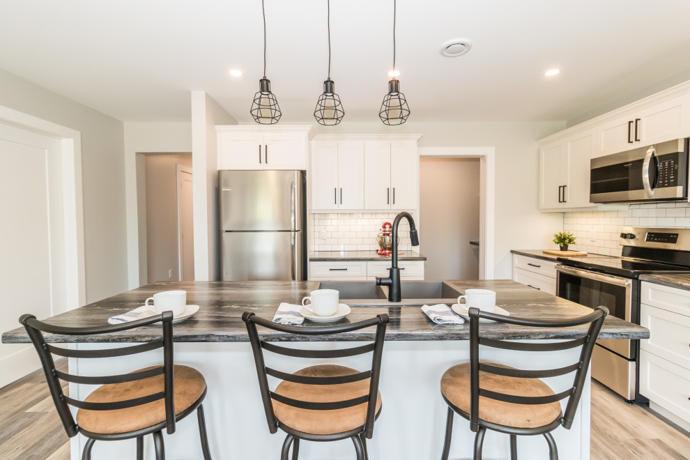 Small l-shaped vinyl floor and brown floor kitchen photo with a drop-in sink, recessed-panel cabinets, white cabinets, laminate countertops, white backsplash, subway tile backsplash, stainless steel appliances, an island and black countertops