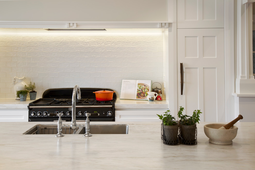 Photo of a traditional galley open plan kitchen in Auckland with a drop-in sink, white cabinets, white splashback, subway tile splashback, black appliances, medium hardwood floors, with island, recessed-panel cabinets and solid surface benchtops.