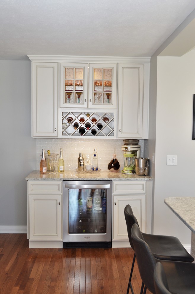 Large traditional l-shaped home bar in Chicago with recessed-panel cabinets, white cabinets, quartzite benchtops, white splashback, glass tile splashback, medium hardwood floors, brown floor and grey benchtop.