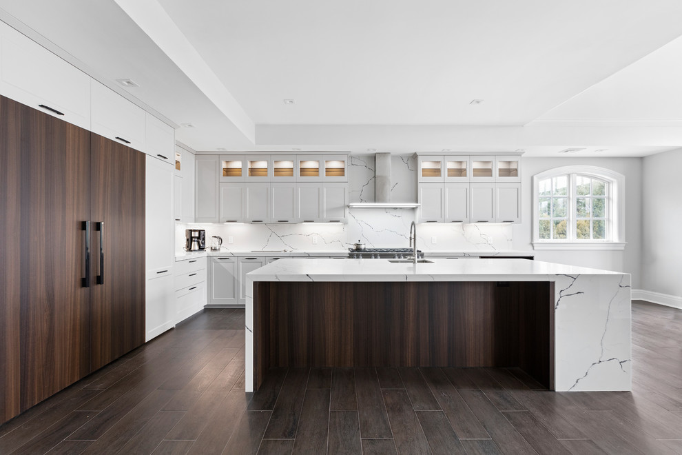 This is an example of an expansive transitional l-shaped open plan kitchen in New York with an undermount sink, recessed-panel cabinets, grey cabinets, quartz benchtops, white splashback, stone slab splashback, stainless steel appliances, dark hardwood floors, with island, brown floor and white benchtop.