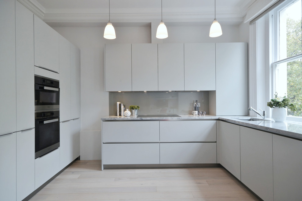 Photo of a contemporary u-shaped kitchen in Gloucestershire with an undermount sink, flat-panel cabinets, white cabinets, grey splashback, stainless steel appliances and light hardwood floors.