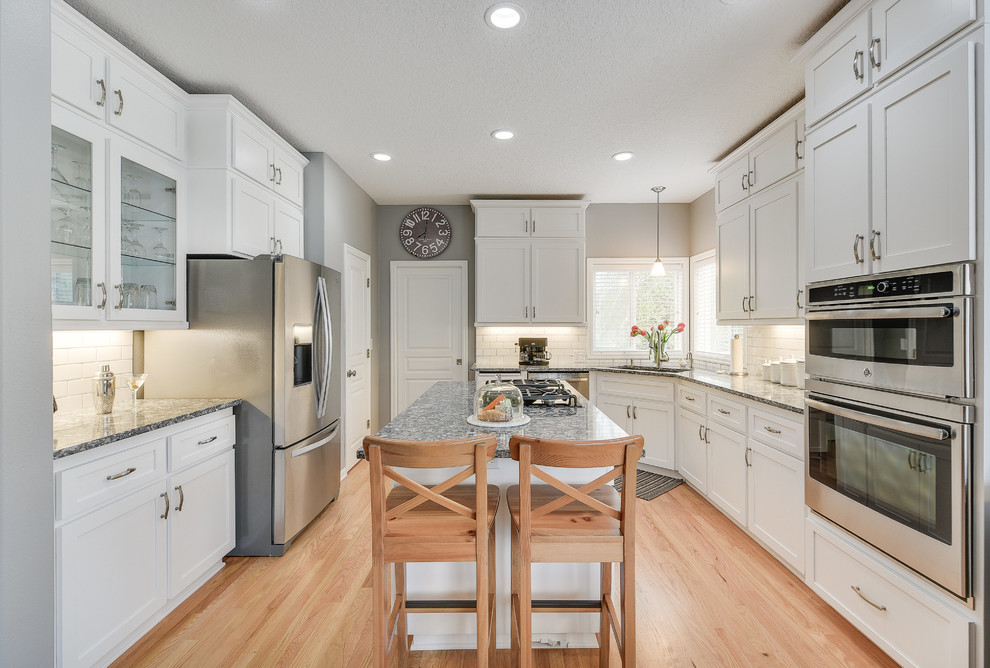 Mid-sized transitional l-shaped light wood floor and brown floor enclosed kitchen photo in Portland with a drop-in sink, shaker cabinets, white cabinets, quartz countertops, white backsplash, subway tile backsplash, an island and stainless steel appliances