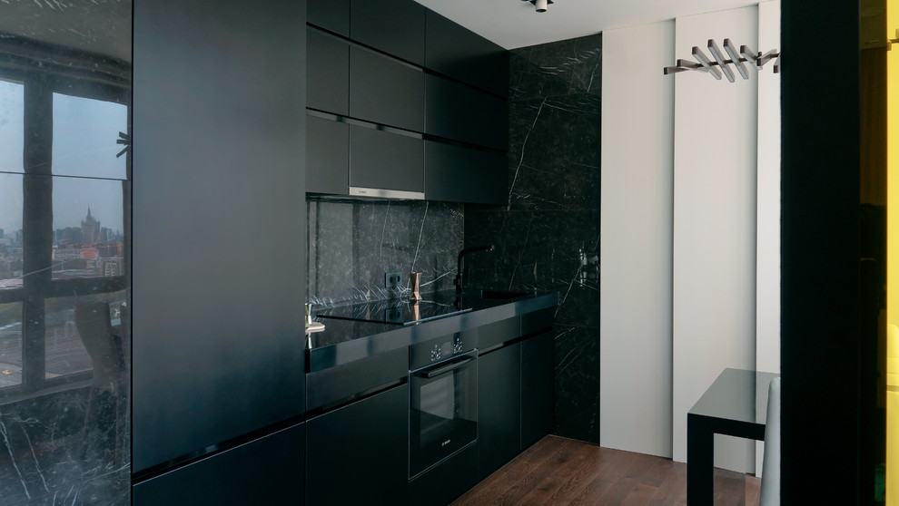 Contemporary single-wall kitchen in Los Angeles with an undermount sink, flat-panel cabinets, black cabinets, black splashback, black appliances, medium hardwood floors, brown floor and black benchtop.
