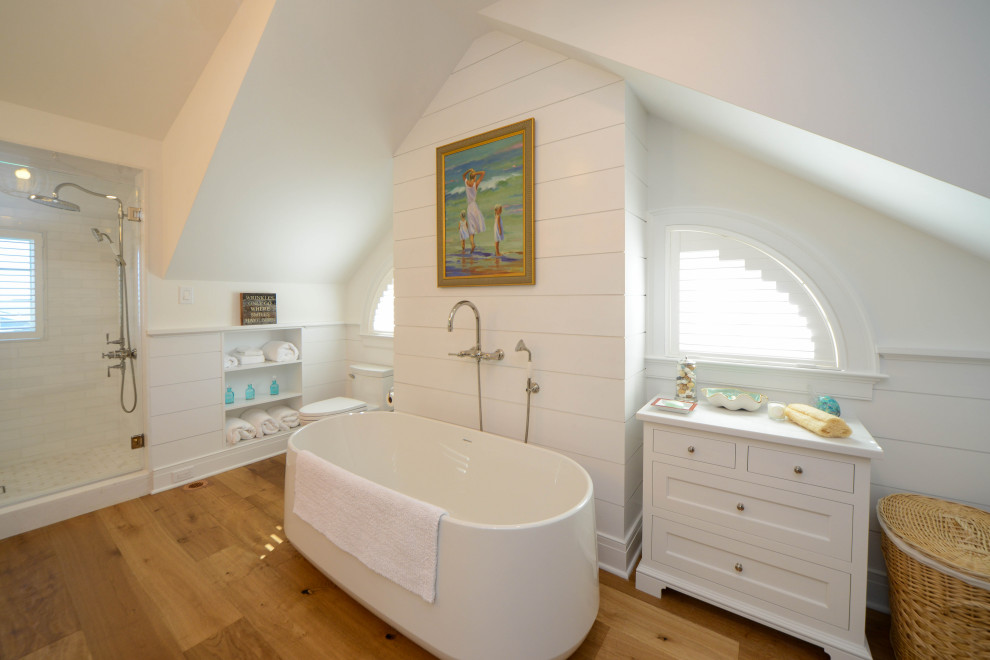 Foto på ett maritimt vit badrum, med ett fristående badkar, en toalettstol med hel cisternkåpa, vit kakel, vita väggar, mellanmörkt trägolv och brunt golv