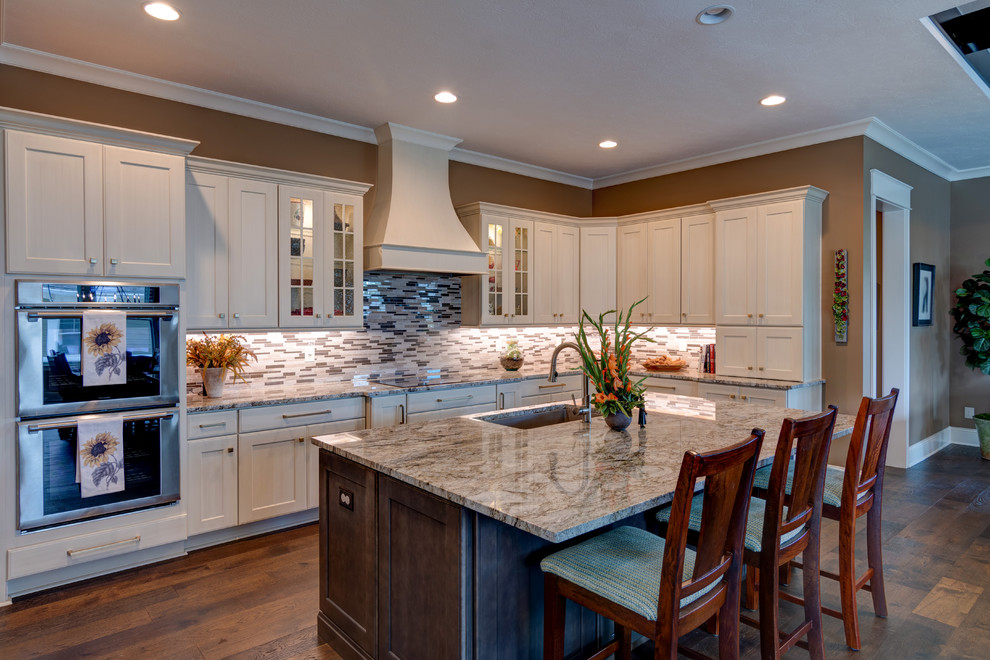 This is an example of a large arts and crafts l-shaped kitchen pantry in Indianapolis with a drop-in sink, shaker cabinets, beige cabinets, granite benchtops, metallic splashback, mosaic tile splashback, panelled appliances, dark hardwood floors, with island, brown floor and beige benchtop.