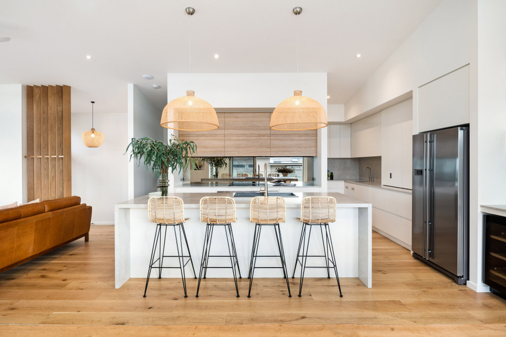 This is an example of a large beach style single-wall kitchen pantry in Wollongong with a double-bowl sink, flat-panel cabinets, white cabinets, metallic splashback, glass sheet splashback, stainless steel appliances, medium hardwood floors, with island, brown floor and beige benchtop.
