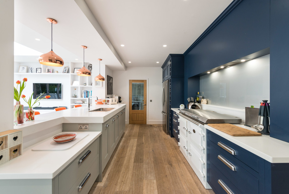 Photo of a mid-sized contemporary galley open plan kitchen in Edinburgh with shaker cabinets, grey splashback, medium hardwood floors, brown floor, white benchtop, an integrated sink, grey cabinets, solid surface benchtops, glass sheet splashback, white appliances and a peninsula.