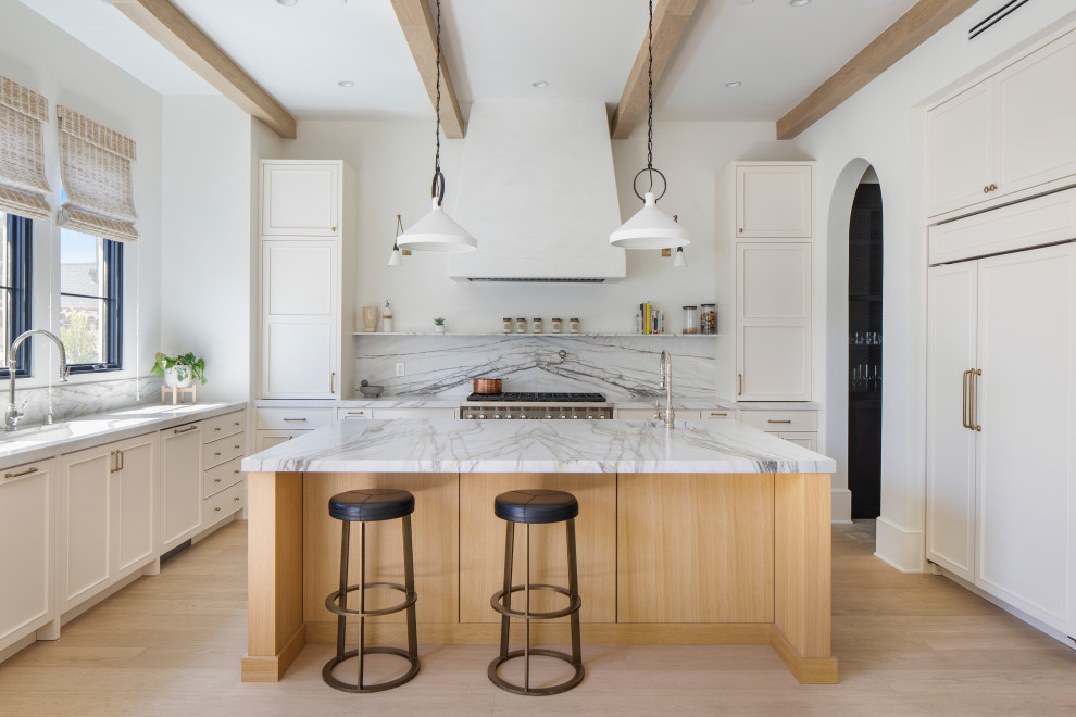 Inspiration for a large mediterranean l-shaped open plan kitchen in Los Angeles with an undermount sink, beaded inset cabinets, white cabinets, marble benchtops, white splashback, marble splashback, stainless steel appliances, light hardwood floors, with island, beige floor, white benchtop and exposed beam.