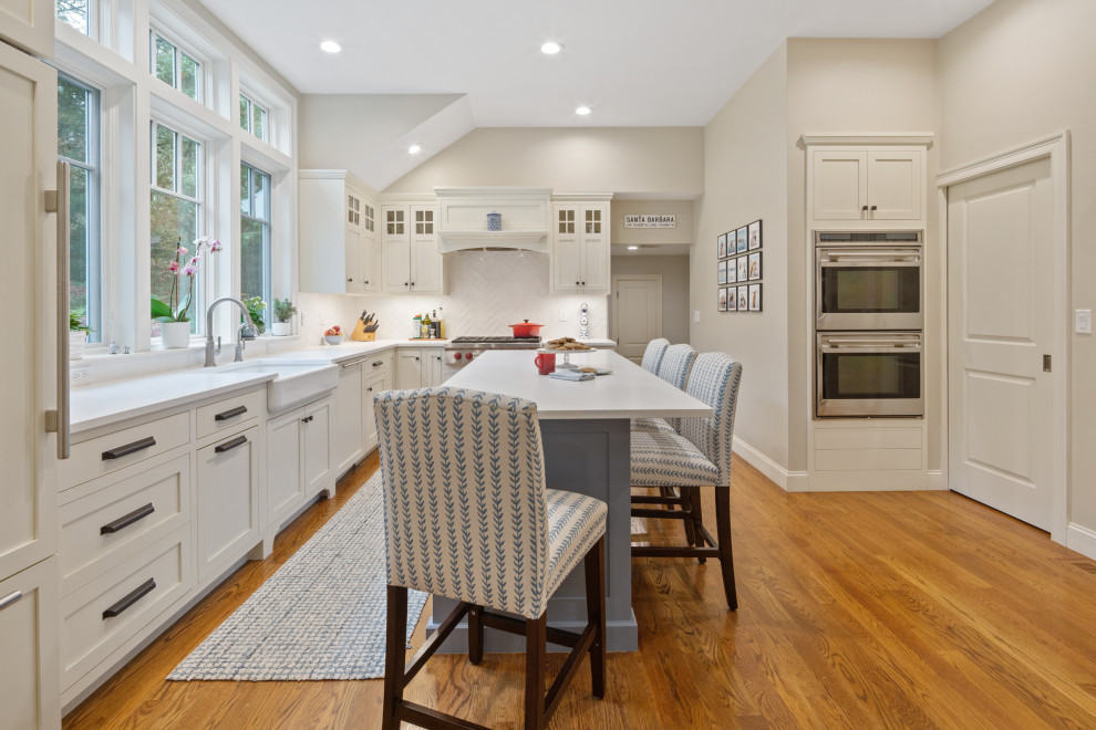 Inspiration for a large coastal l-shaped medium tone wood floor and brown floor kitchen pantry remodel in Boston with a farmhouse sink, shaker cabinets, white cabinets, quartz countertops, white backsplash, ceramic backsplash, stainless steel appliances, an island and white countertops