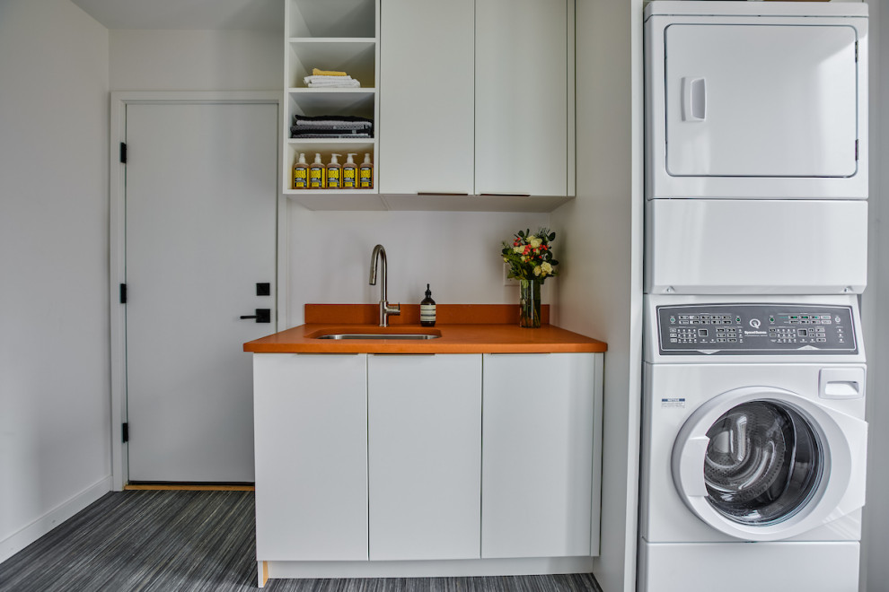 デトロイトにある高級な中くらいなミッドセンチュリースタイルのおしゃれな家事室 (アンダーカウンターシンク、フラットパネル扉のキャビネット、クオーツストーンカウンター、白いキッチンパネル、白い壁、リノリウムの床、上下配置の洗濯機・乾燥機、グレーの床、ll型、オレンジのキッチンカウンター、白いキャビネット) の写真