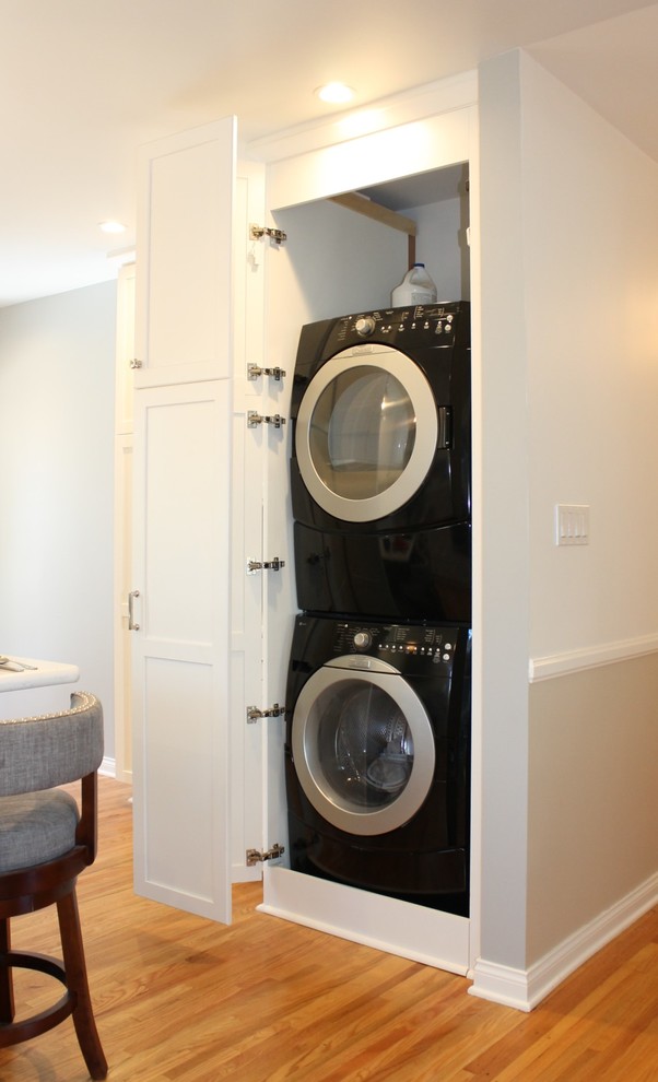 Photo of a mid-sized transitional single-wall laundry cupboard in Los Angeles with shaker cabinets, white cabinets, quartz benchtops, grey walls, medium hardwood floors, a stacked washer and dryer, brown floor and white benchtop.