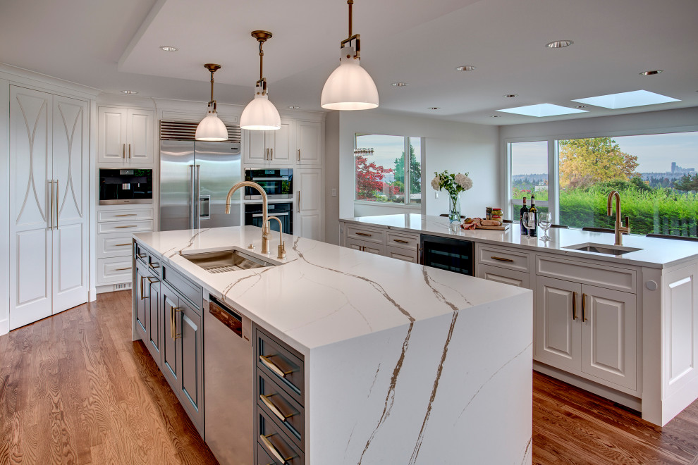 Inspiration for a large transitional l-shaped open plan kitchen in Seattle with a single-bowl sink, raised-panel cabinets, white cabinets, quartz benchtops, white splashback, engineered quartz splashback, stainless steel appliances, medium hardwood floors, multiple islands, brown floor, white benchtop and recessed.