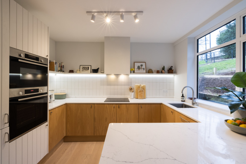 Inspiration for a mid-sized contemporary u-shaped eat-in kitchen in London with an undermount sink, louvered cabinets, light wood cabinets, quartzite benchtops, white splashback, ceramic splashback, stainless steel appliances, light hardwood floors, a peninsula, brown floor and white benchtop.