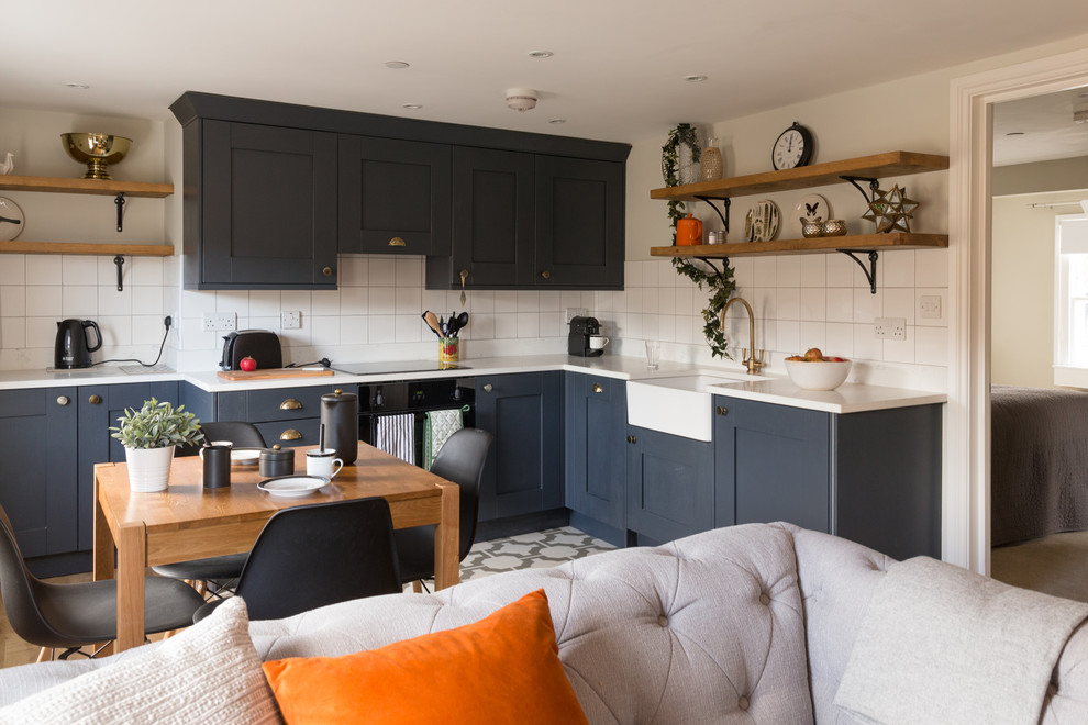 Photo of a mid-sized eclectic l-shaped open plan kitchen in Sussex with a farmhouse sink, shaker cabinets, black cabinets, solid surface benchtops, white splashback, ceramic splashback, black appliances, vinyl floors and no island.