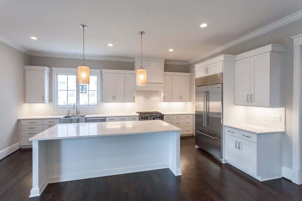 This is an example of an arts and crafts l-shaped open plan kitchen in DC Metro with a farmhouse sink, recessed-panel cabinets, white cabinets, white splashback, subway tile splashback, stainless steel appliances, dark hardwood floors, with island, brown floor and white benchtop.