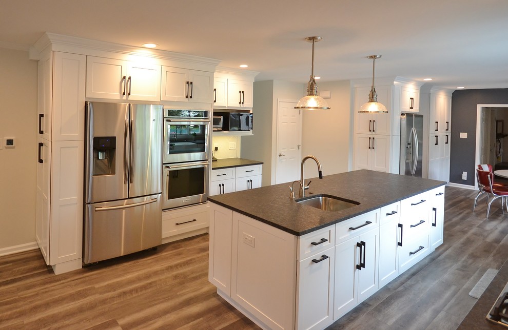 This is an example of a large transitional l-shaped eat-in kitchen in Philadelphia with a farmhouse sink, recessed-panel cabinets, white cabinets, granite benchtops, white splashback, subway tile splashback, stainless steel appliances, vinyl floors, with island and brown floor.