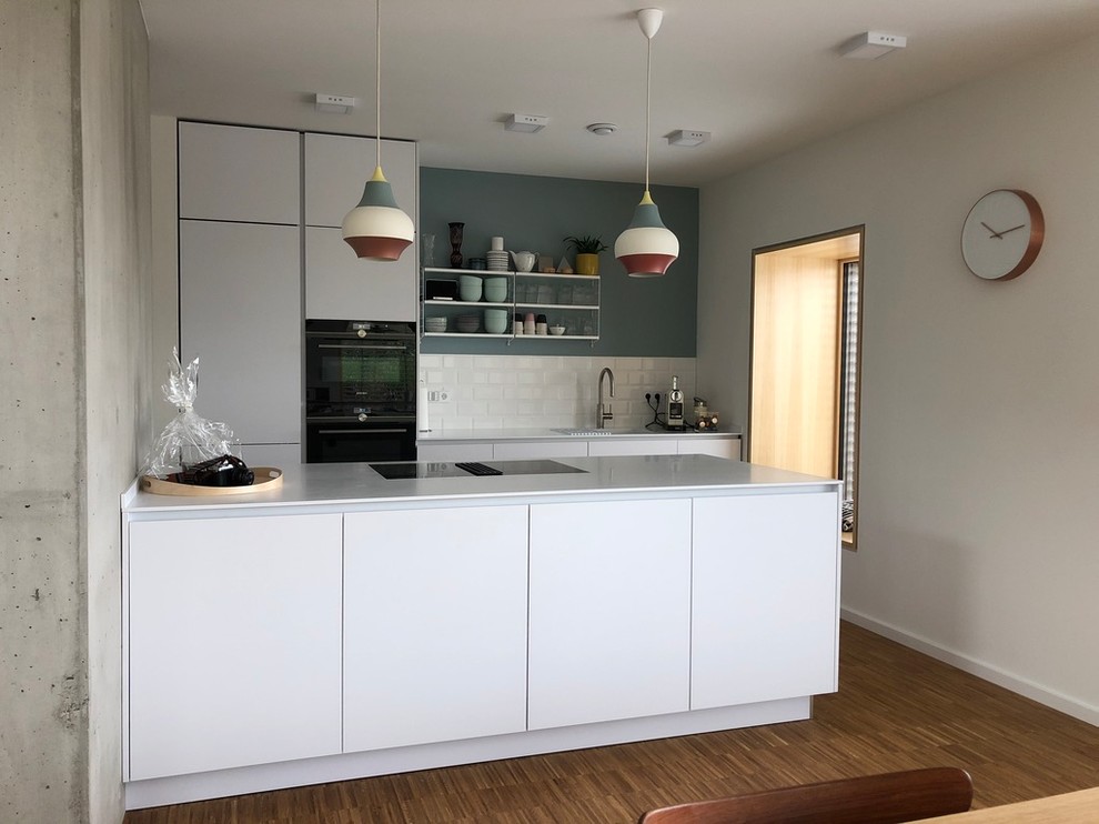 Zweizeilige, Mittelgroße, Offene Nordische Küche mit integriertem Waschbecken, weißen Schränken, Mineralwerkstoff-Arbeitsplatte, Rückwand aus Metrofliesen, schwarzen Elektrogeräten, braunem Holzboden, braunem Boden, weißer Arbeitsplatte, flächenbündigen Schrankfronten, Küchenrückwand in Weiß und Halbinsel in München
