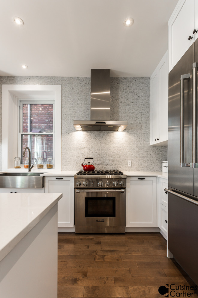 Example of a mid-sized transitional l-shaped medium tone wood floor eat-in kitchen design in Montreal with a farmhouse sink, shaker cabinets, white cabinets, quartzite countertops, white backsplash, mosaic tile backsplash, stainless steel appliances and an island