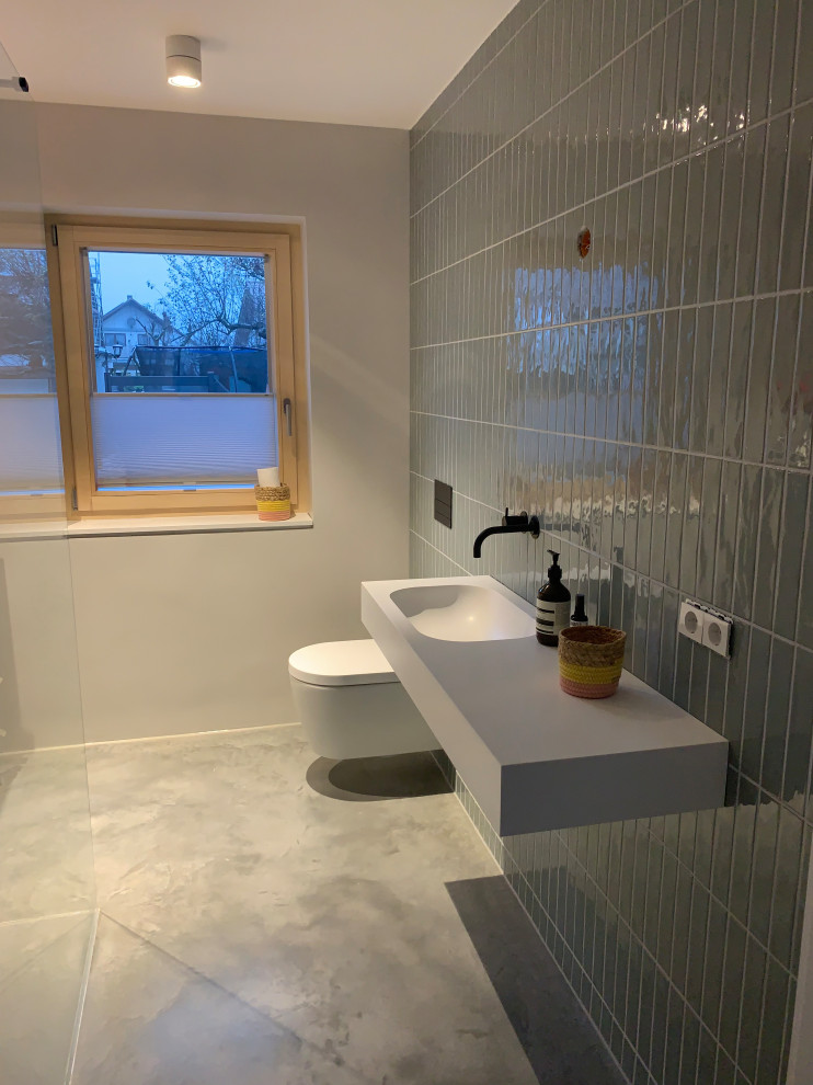 Photo of a mid-sized contemporary 3/4 bathroom in Stuttgart with a curbless shower, a two-piece toilet, green tile, matchstick tile, a wall-mount sink, solid surface benchtops, an open shower, white benchtops, a single vanity, a floating vanity, green walls, concrete floors, grey floor and wallpaper.