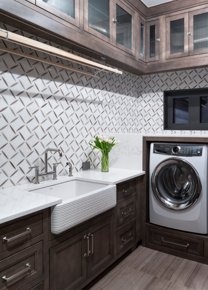 Example of a mid-sized transitional u-shaped porcelain tile and brown floor dedicated laundry room design in Denver with a farmhouse sink, beaded inset cabinets, medium tone wood cabinets, quartz countertops, white walls, an integrated washer/dryer and white countertops